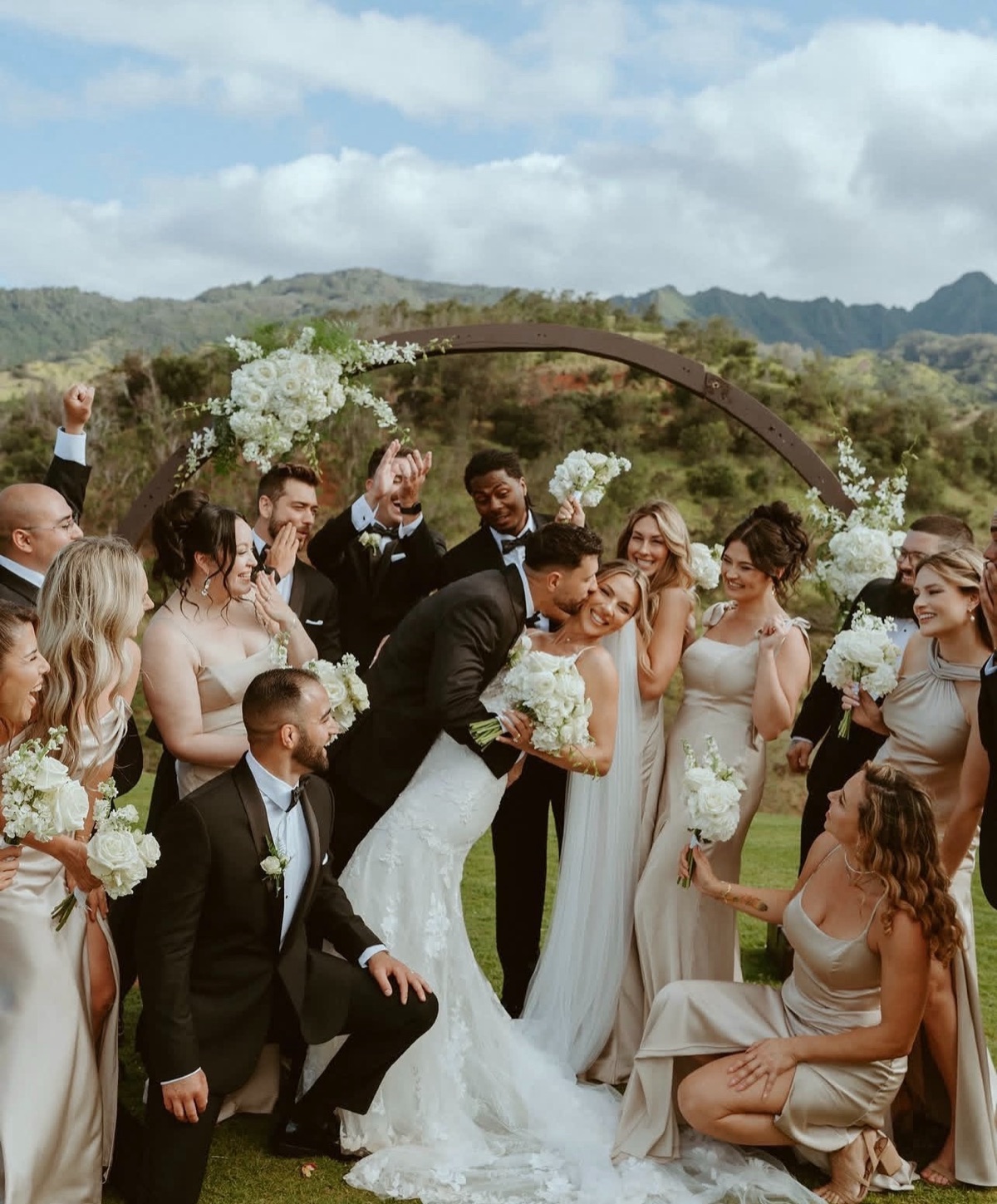 A wedding party laughing under a floral arch with mountains behind them